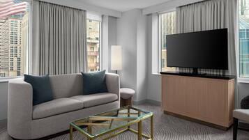 A modern hotel room with a sofa, a glass coffee table, a flat-screen TV, and large windows with curtains.