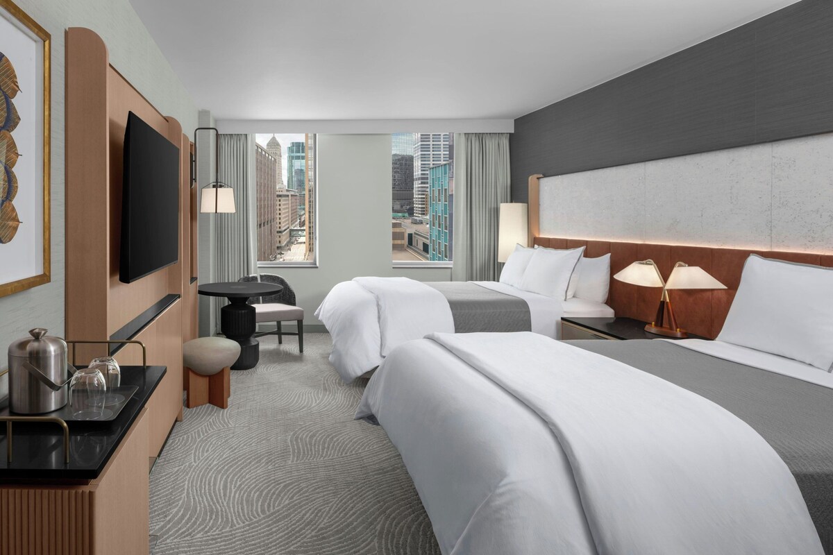 Ein Hotelzimmer mit zwei Betten, einem Fernseher, einem kleinen Tisch und Blick auf die Stadt.