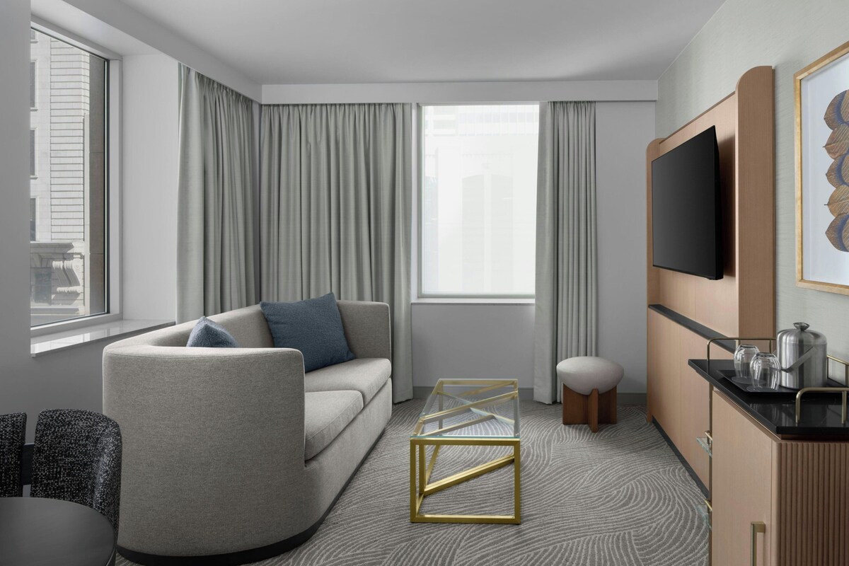 Ein modernes Hotelzimmer mit einer Couch, einem Glastisch, einem Flachbildfernseher, einer Essecke und Blick auf die Stadt.