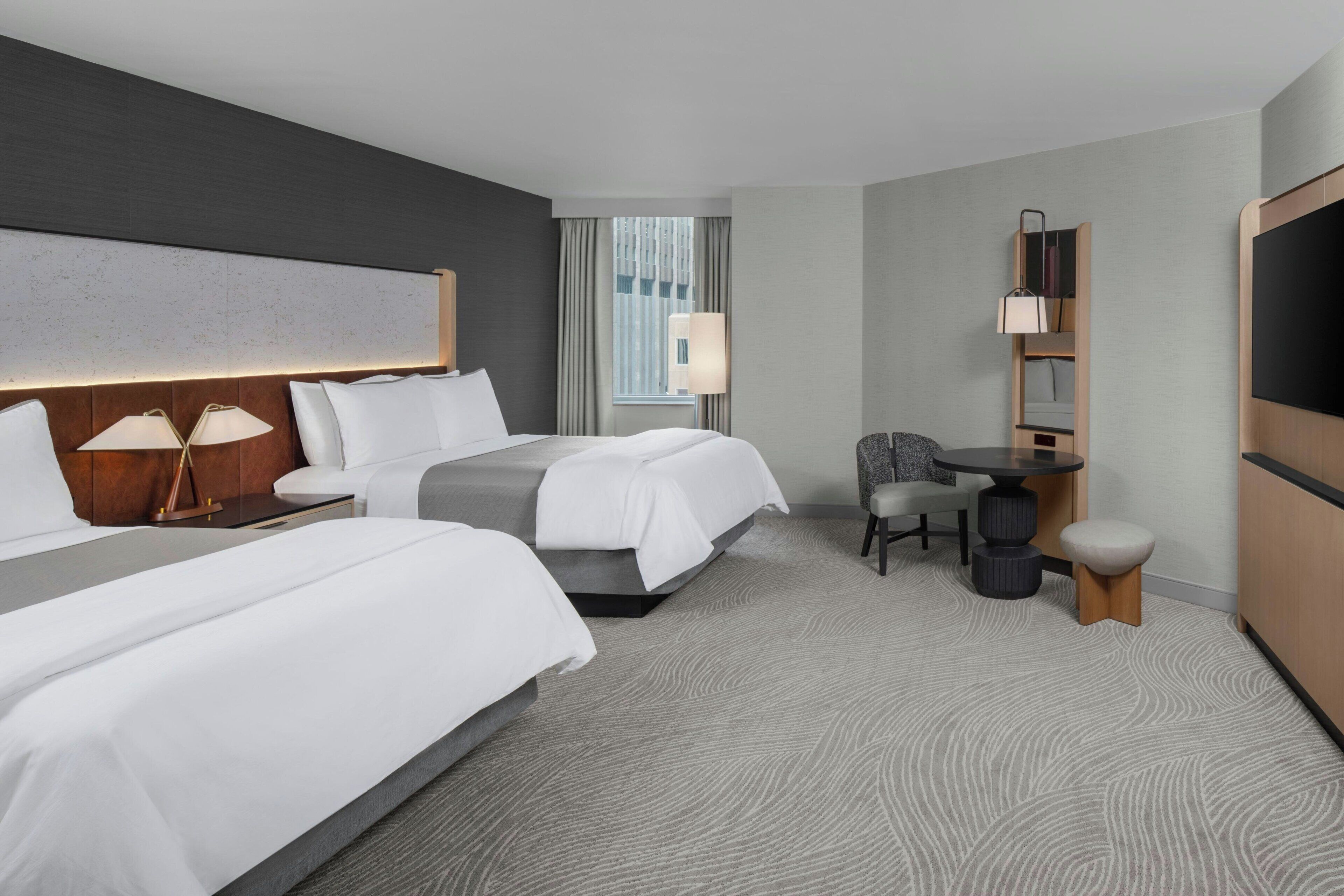Quarto de hotel com duas camas, uma escrivaninha, uma cadeira, televisão e janela com cortinas.