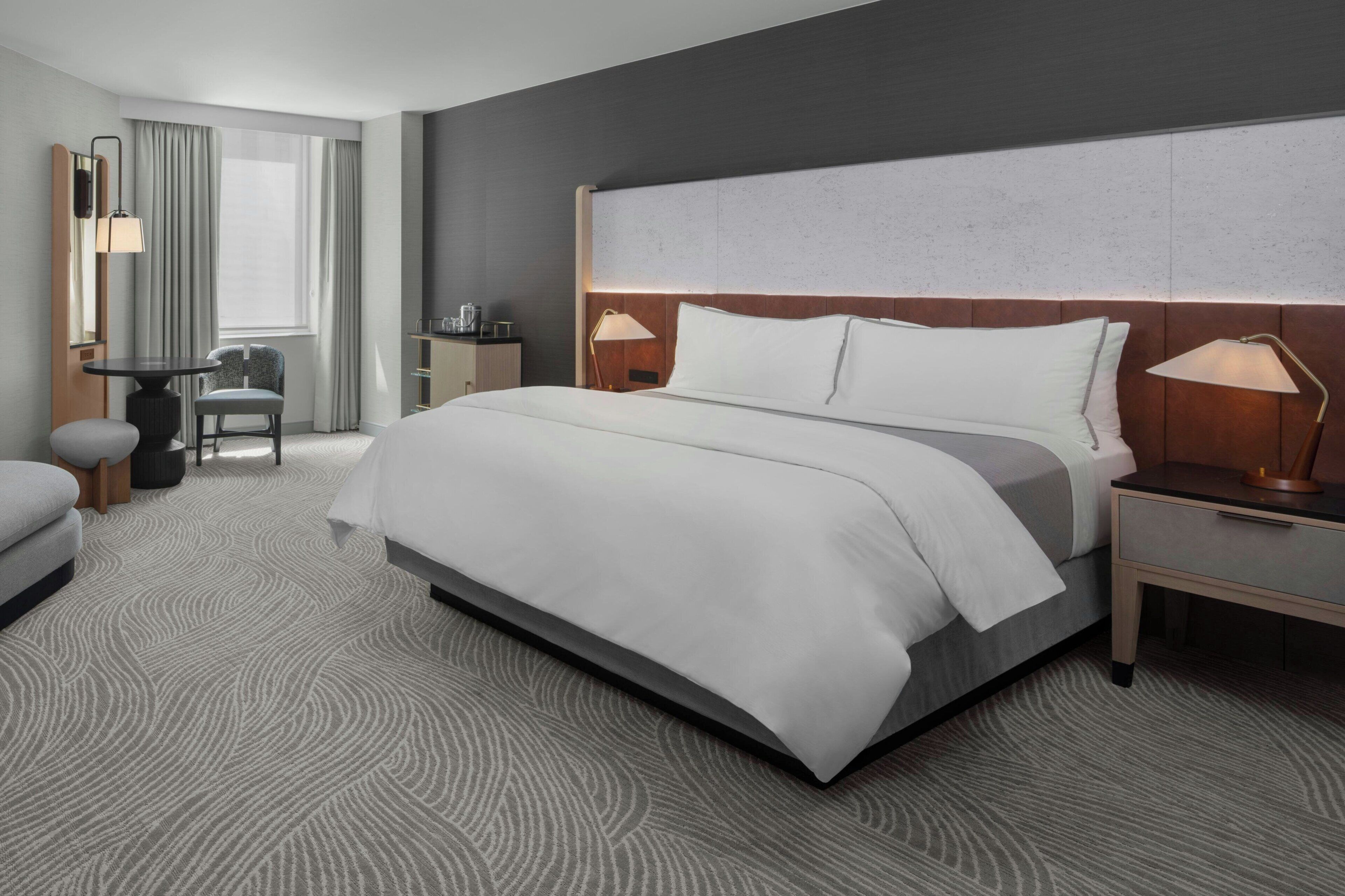 Habitación de hotel con una cama grande, un escritorio con silla, una lámpara y un ventanal con cortinas.