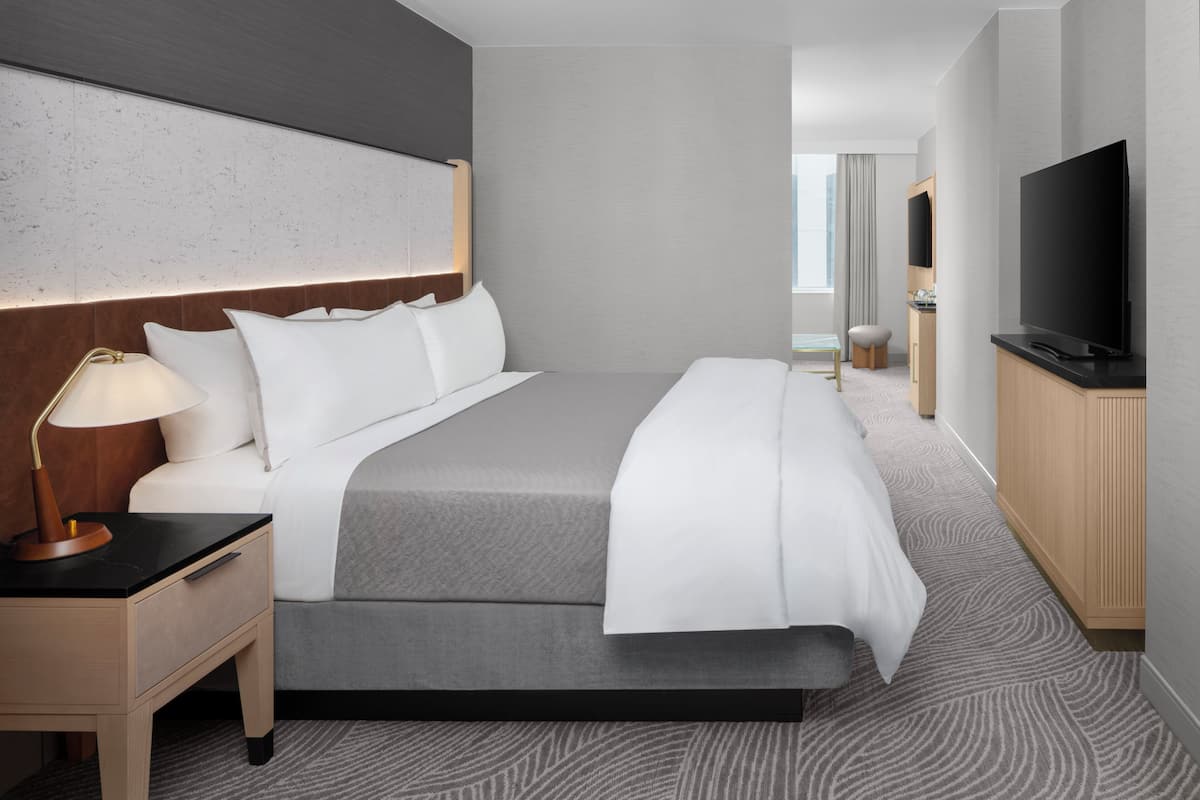 A modern hotel room with a large bed, a TV, a desk with a lamp, and a view of the hallway.