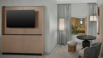 Quarto de hotel com televisão de tela plana fixada em um painel de madeira, uma mesa de jantar redonda com duas cadeiras, um pequeno banquinho e uma grande janela com cortinas.