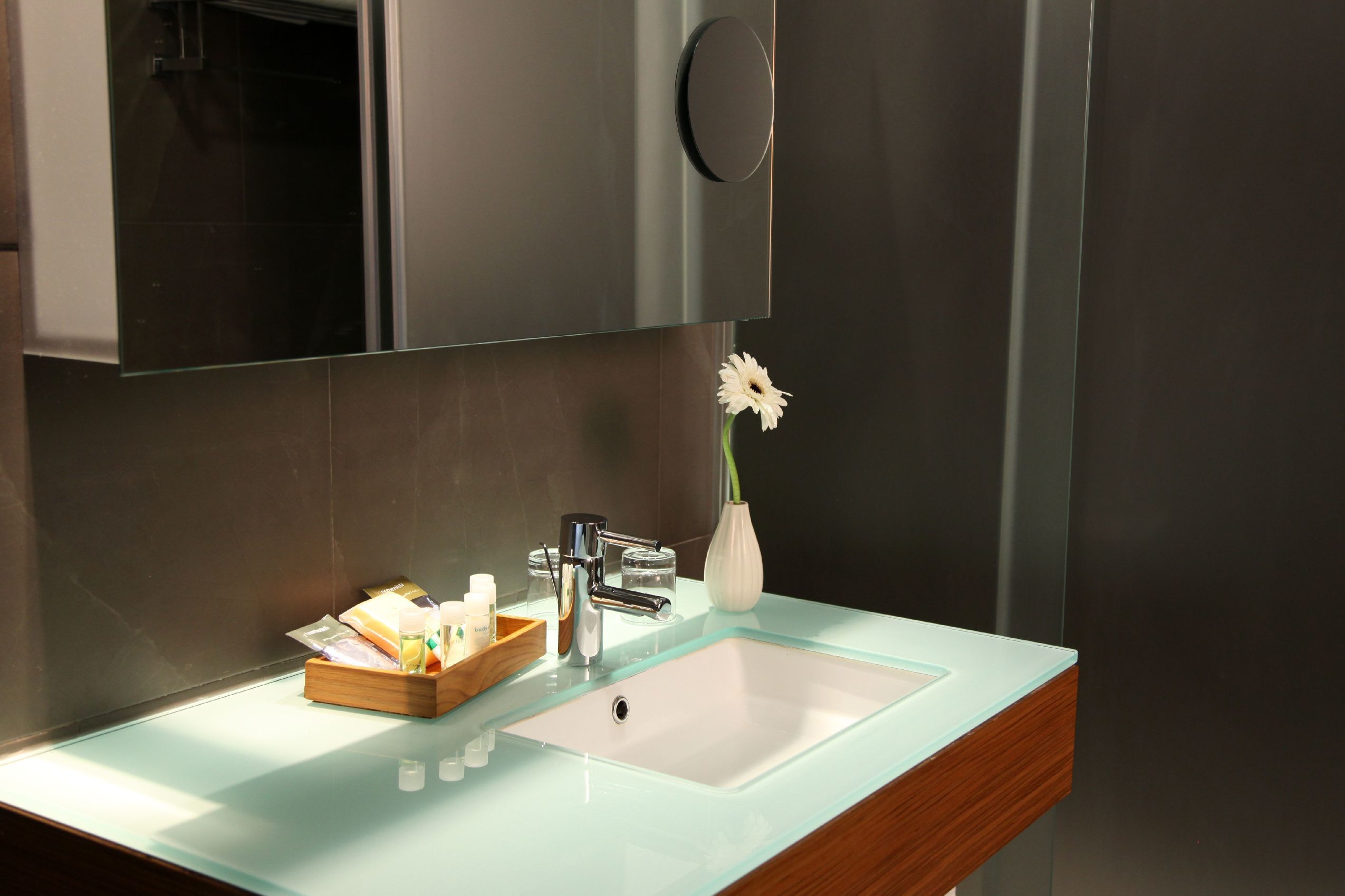 A modern bathroom with a glass sink, a wooden tray, a vase with a flower, and a mirror above the sink.