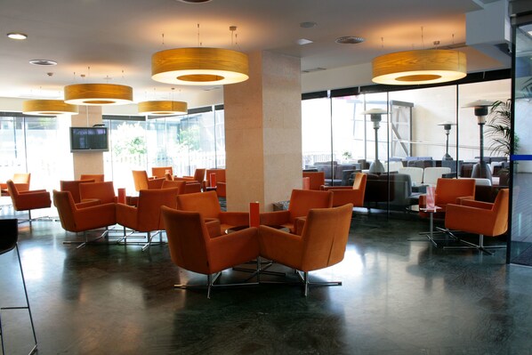 A spacious lobby with modern orange chairs, large wooden pendant lights, and a large TV screen mounted on a pillar.