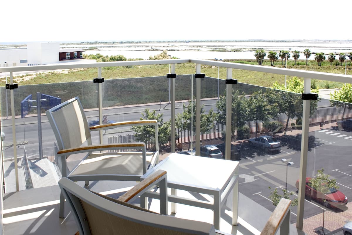 A balcony with white chairs and a table, overlooking a parking area with cars and a river.