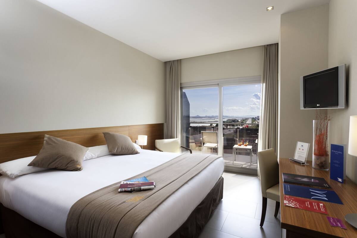 A hotel room with a large bed, a desk, a TV, and a balcony with a view.