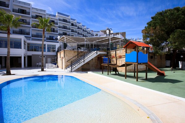 A swimming pool with a slide and a play structure, surrounded by a building with balconies and a clear sky. - Palmanova Suites by TRH (Calvia)
