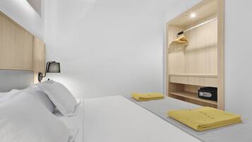 Une chambre d’hôtel moderne avec un lit bien fait, une table de chevet avec une lampe, une armoire intégrée avec un miroir et une petite étagère avec une radio.