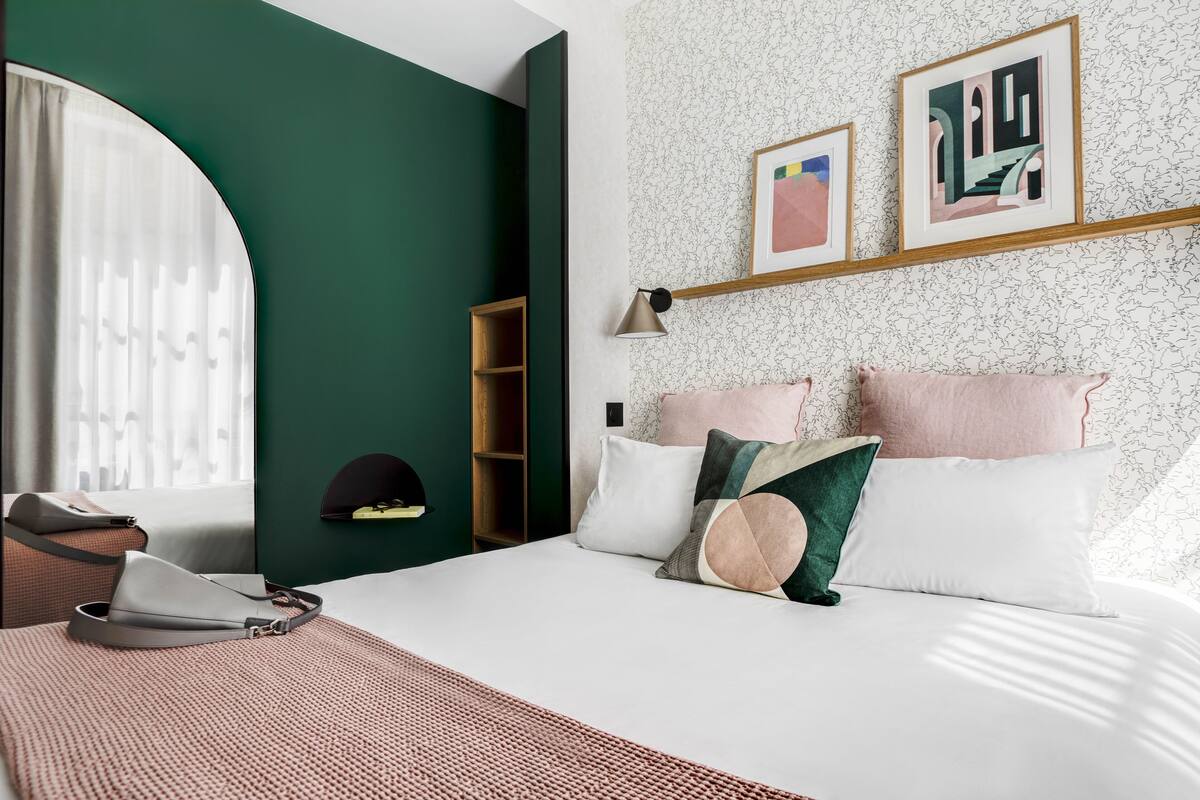 A bedroom with a bed, pillows, a bedside table, a lamp, a wardrobe, and a window with curtains.