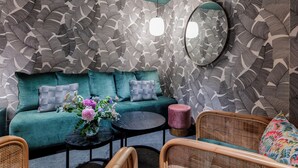 A room with a patterned wall, a teal sofa with cushions, a round table with a vase of flowers, wicker chairs, and a round mirror. - Republik Hôtel (Lyon)