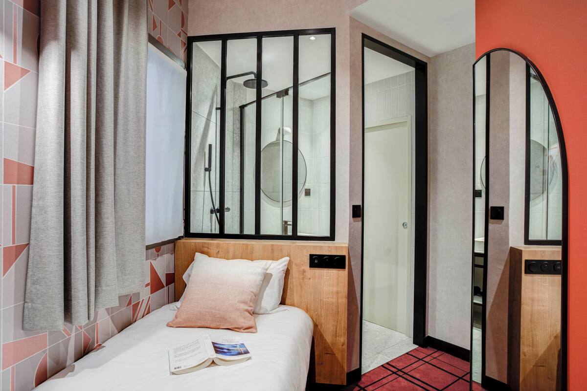 A hotel room with a bed, a mirror, a shower area, and a door leading to another room.