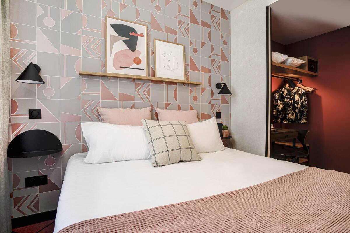 A neatly arranged bedroom with a bed, pillows, a bedside table, a lamp, and a shelf with framed pictures.