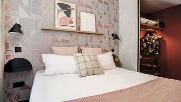 A neatly arranged bedroom with a bed, pillows, a bedside table, a lamp, and a shelf with framed pictures.