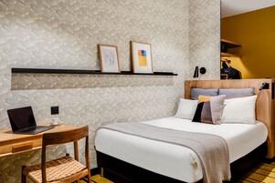 Camera d'albergo ordinata con un letto, una scrivania con un portatile, una sedia e uno scaffale con due quadri incorniciati.