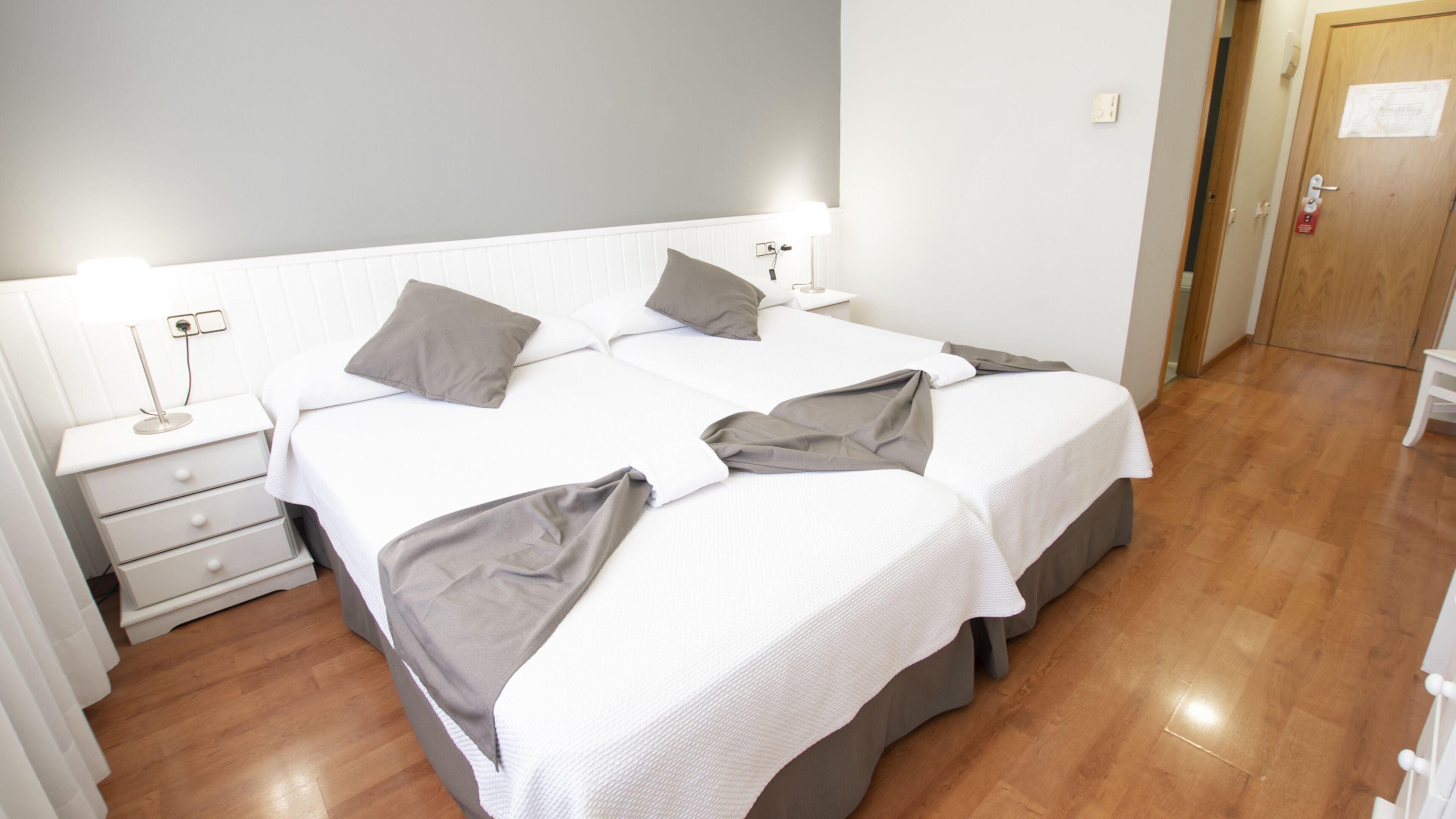 A hotel room with two beds, a bedside table with a lamp, and a wooden floor.