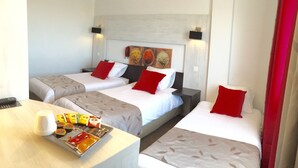 A hotel room with three beds, each with a red pillow, a bedside table with a tray of snacks, and a wall-mounted TV. - Victoria Hotel (Perpignan)