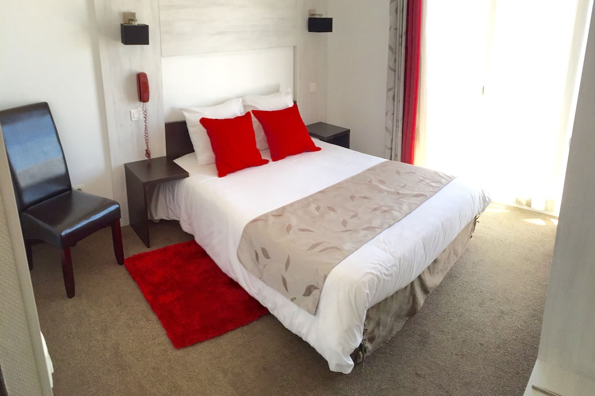A hotel room with a bed, a red rug, a chair, and a nightstand with a phone.
