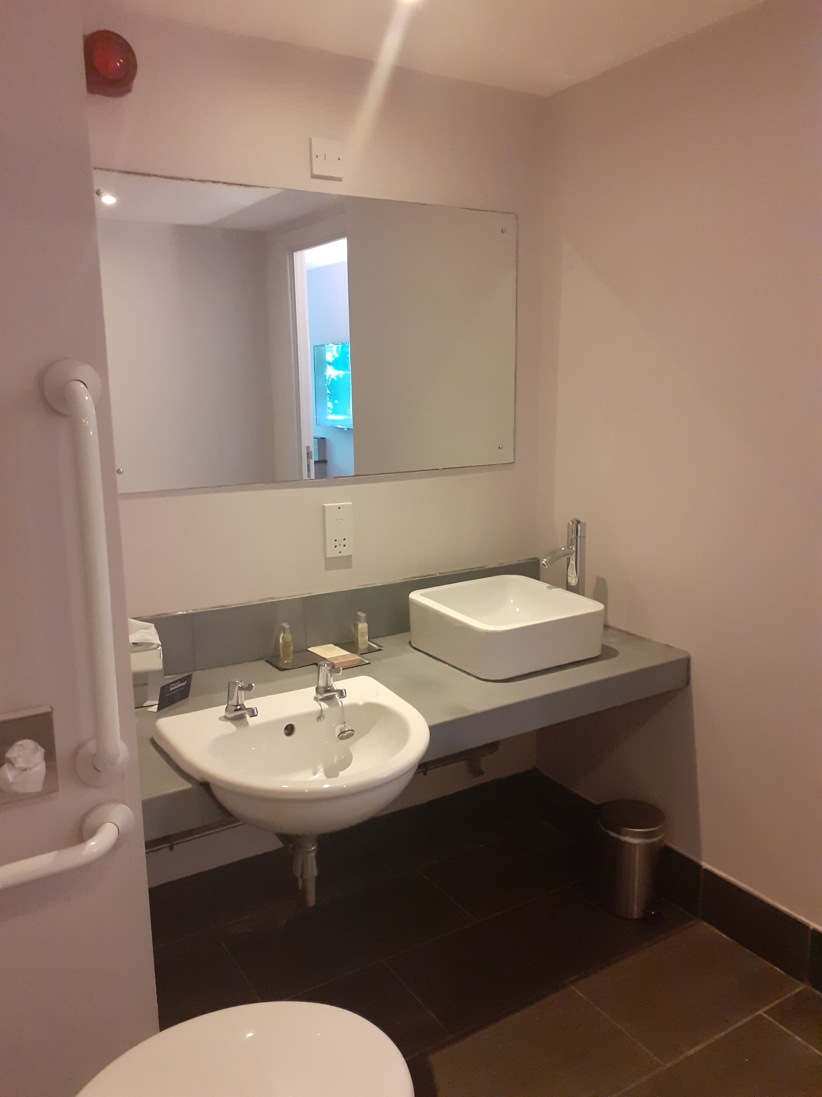 A bathroom with two sinks, a mirror, a handrail, and a trash can.