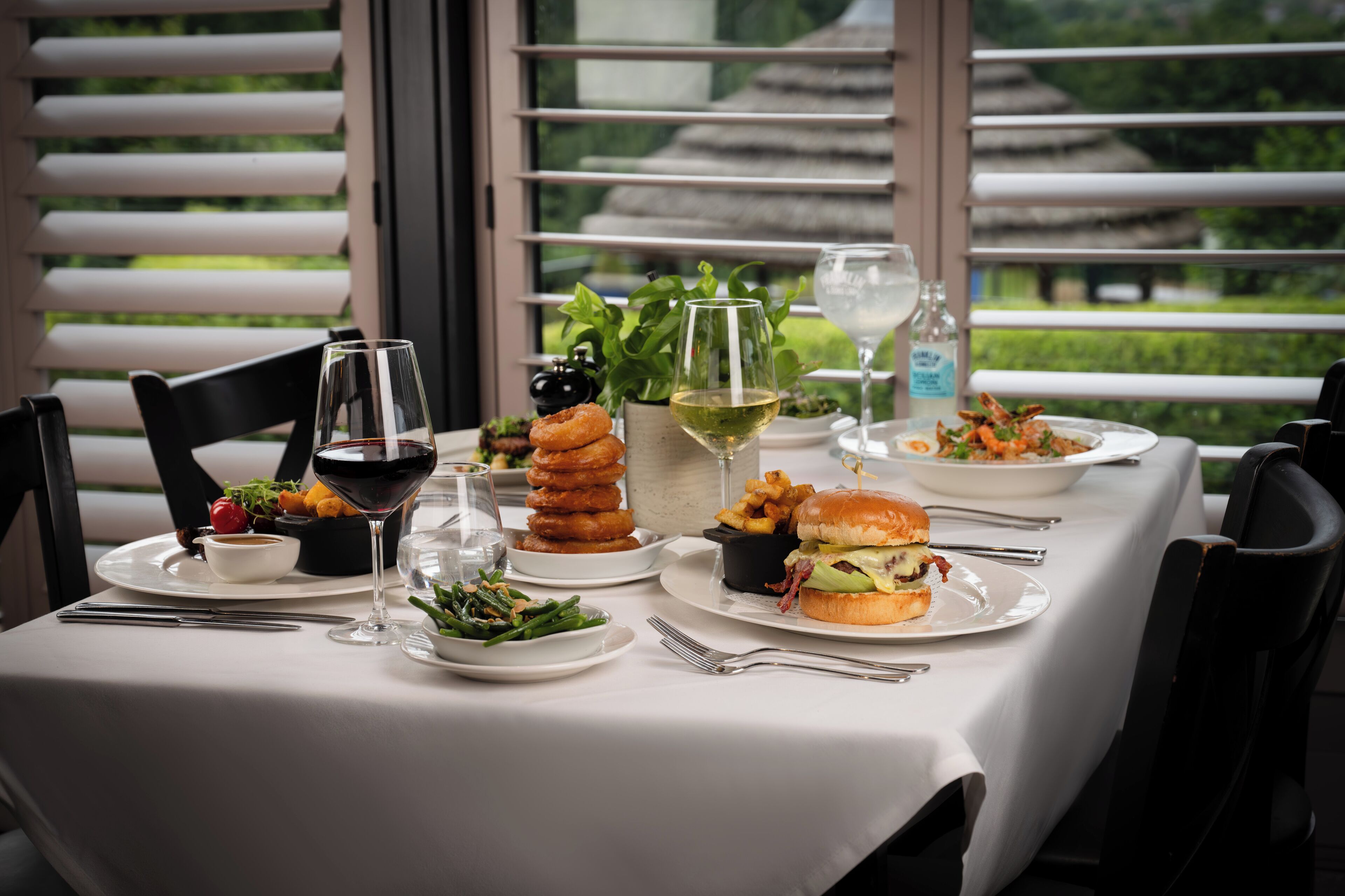 A dining table set with various dishes, glasses, and cutlery, with a view of greenery and traditional architecture through the window.
