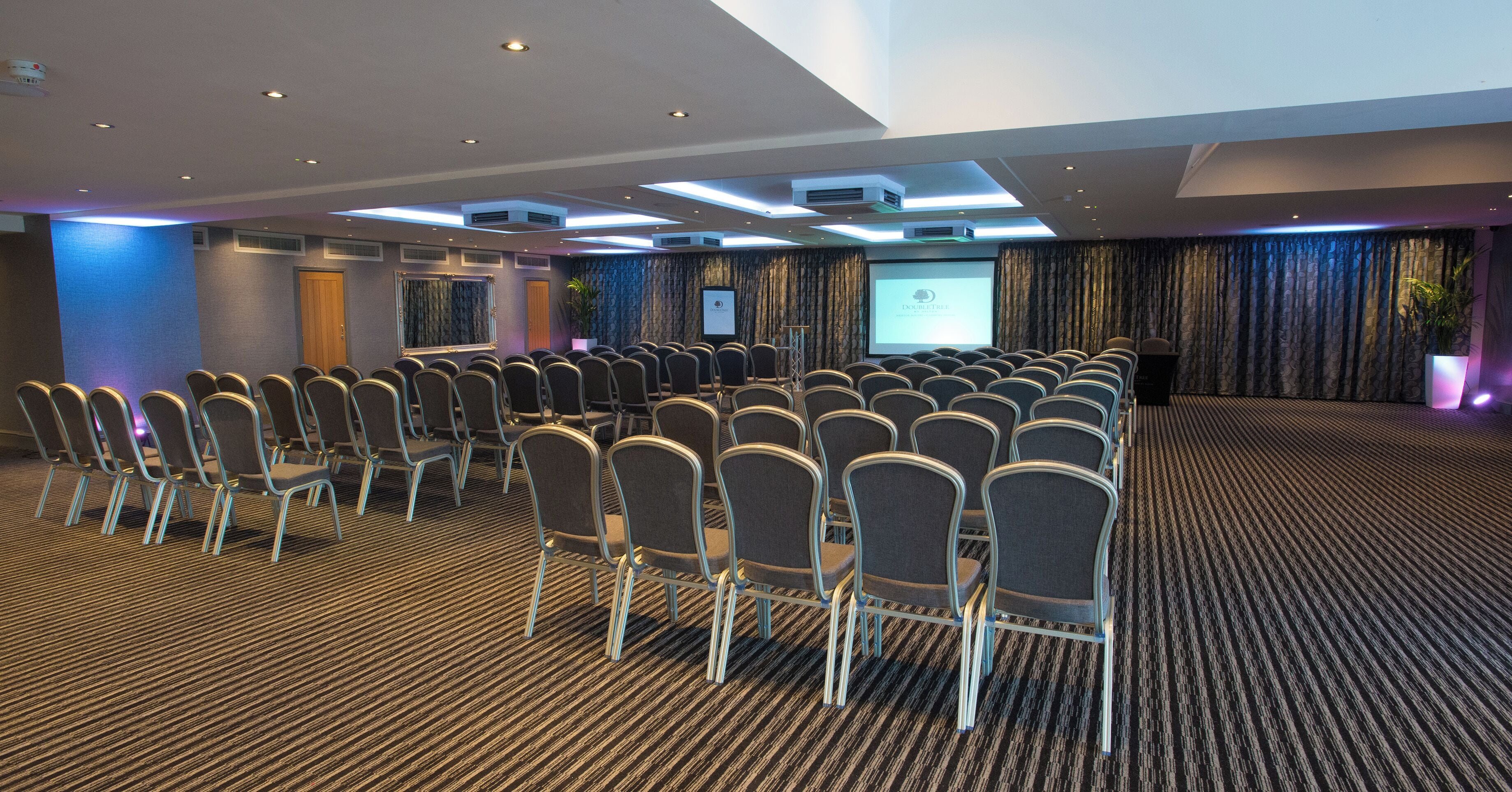 A conference room with rows of chairs, a projector screen, and a carpeted floor.