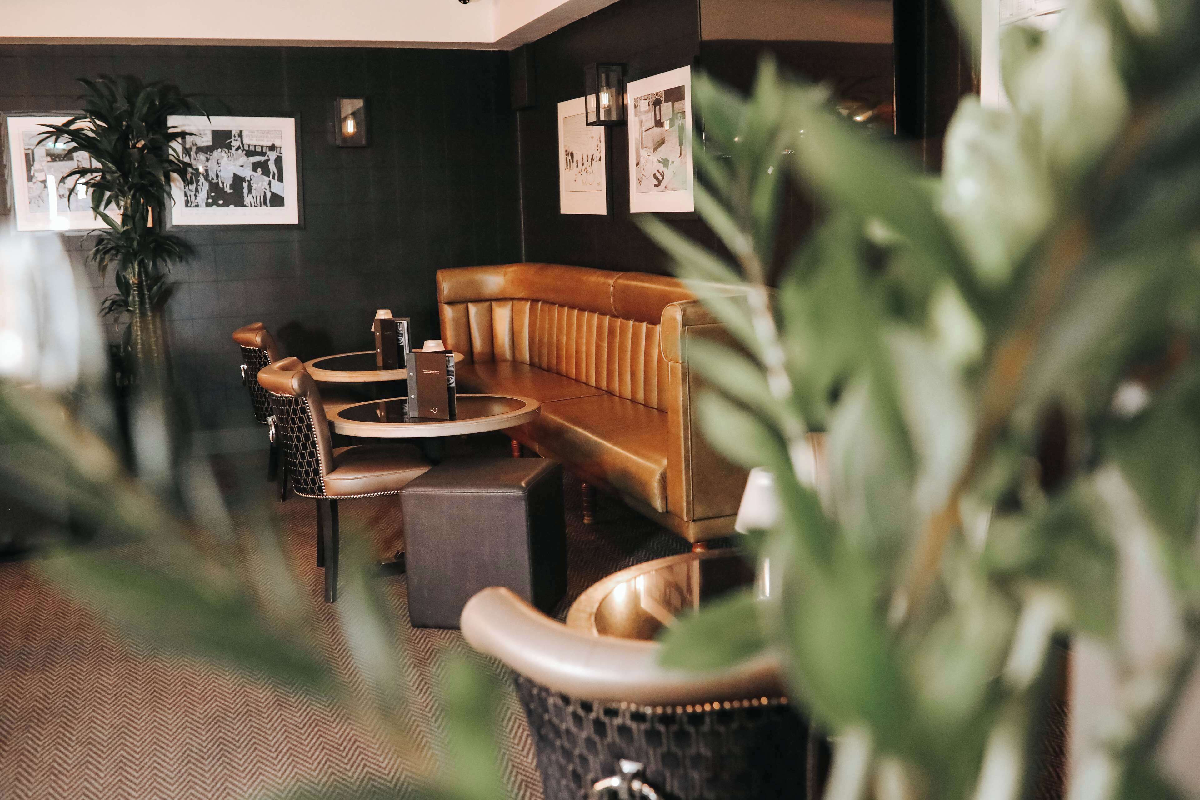 A cozy indoor seating area with a leather sofa, a round table, and a small round table. There are framed pictures on the wall and a potted plant in the foreground.