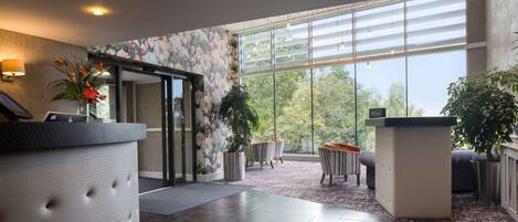A modern hotel lobby with a reception desk, seating area, and large windows offering a view of the outdoors.