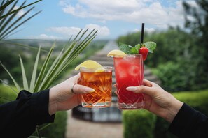Dos manos sostienen vasos con bebidas heladas, uno con una rodaja de limón y menta, y el otro con una fresa y menta, sobre un fondo de vegetación y cielo despejado.