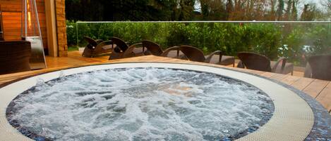 Un jacuzzi con detalle de mosaico, rodeado de sillones de mimbre, sobre una terraza de madera con vista a follaje.