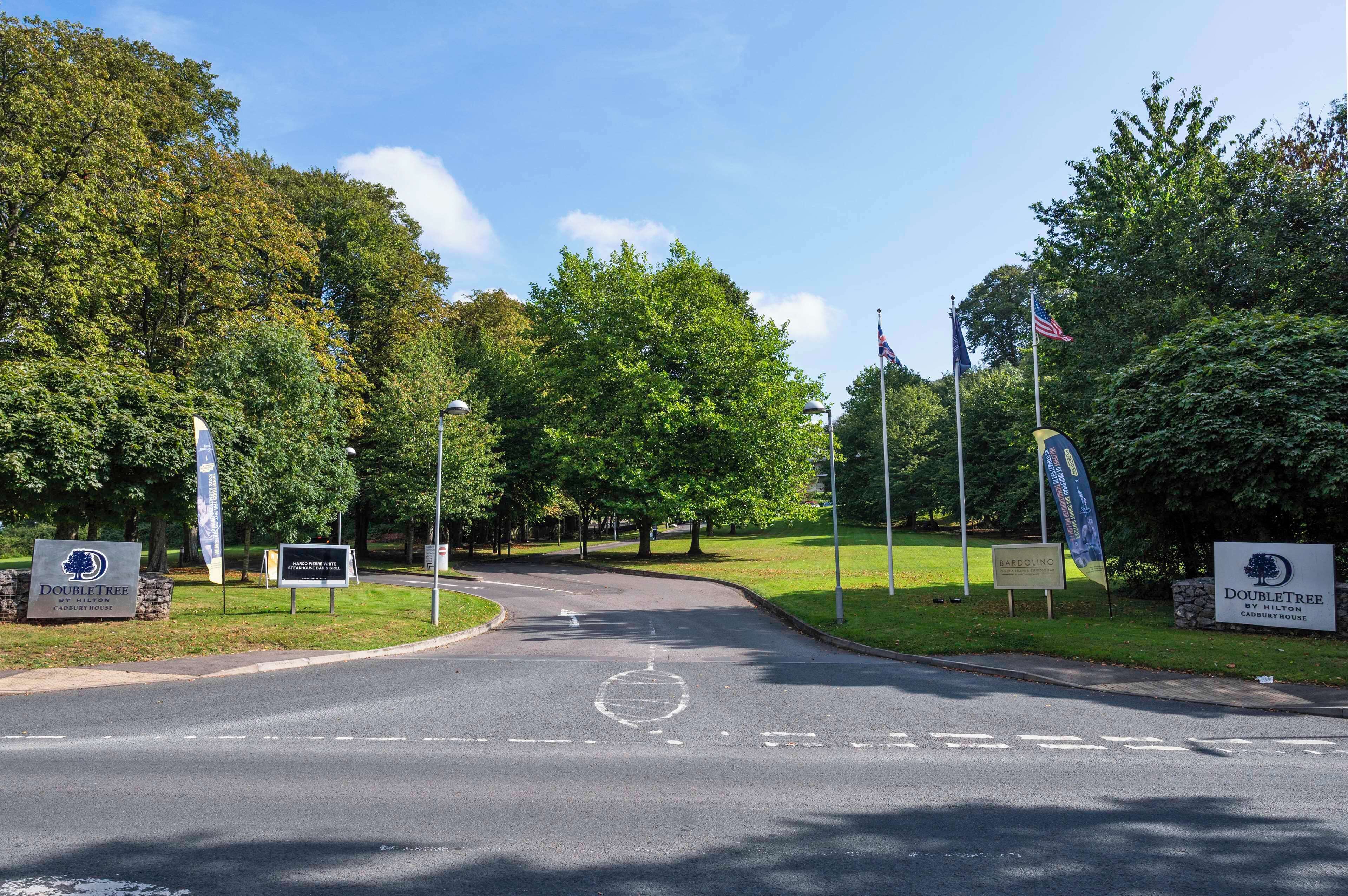 A road leading to a gated entrance with a sign for DoubleTree by Hilton Cary, surrounded by lush greenery and flags.