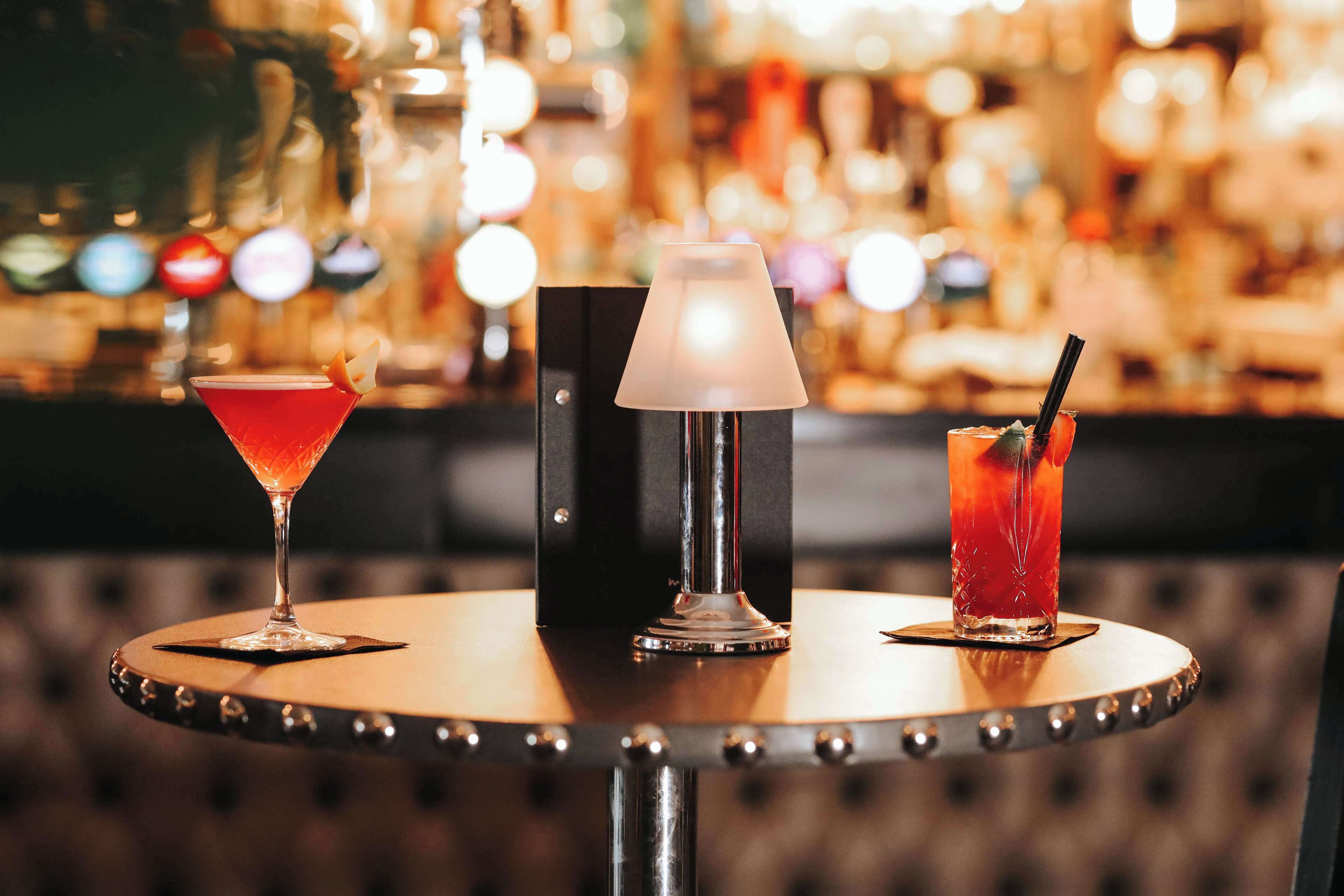 A bar setting with three drinks on a round table, a lamp, and a blurred background of a bar.