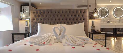 Une chambre d’hôtel moderne avec une tête de lit capitonnée, un lit décoré de pétales de rose et des serviettes disposées en forme de cœur, une coiffeuse avec un miroir et un plancher en bois.