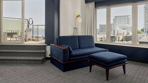 A modern hotel room with a sofa, ottoman, and a view of the city through large windows. - Courtyard by Marriott Halifax Downtown (Halifax)