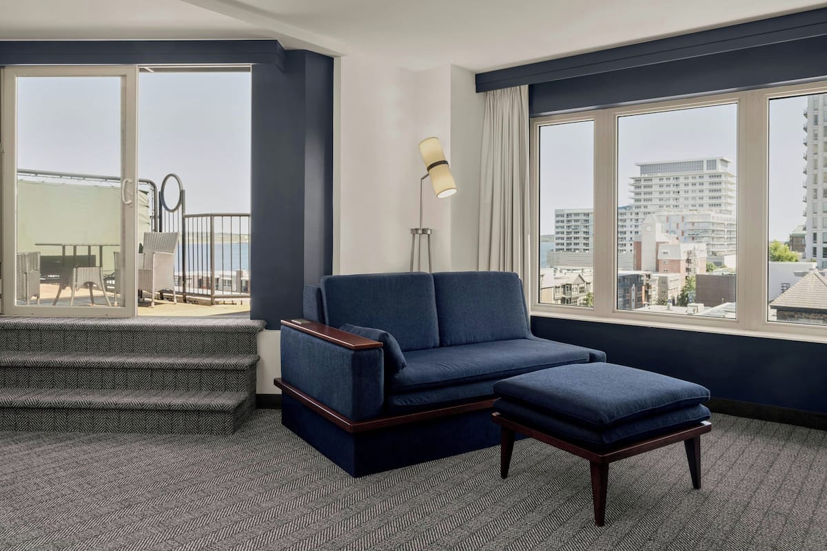 A modern hotel room with a sofa, ottoman, and a view of the city through large windows.