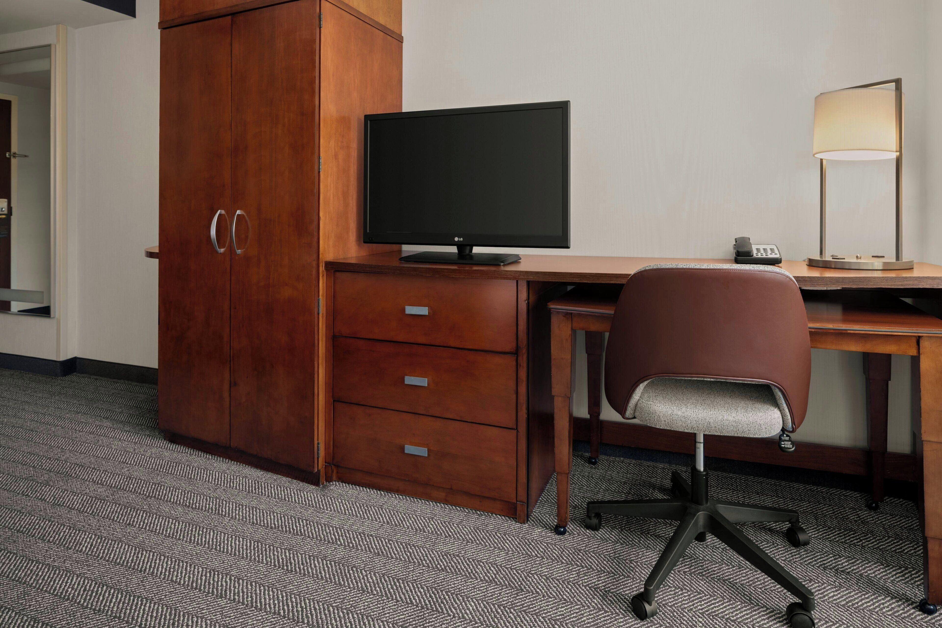 A hotel room with a wooden desk, a computer monitor, a lamp, a telephone, and a chair.