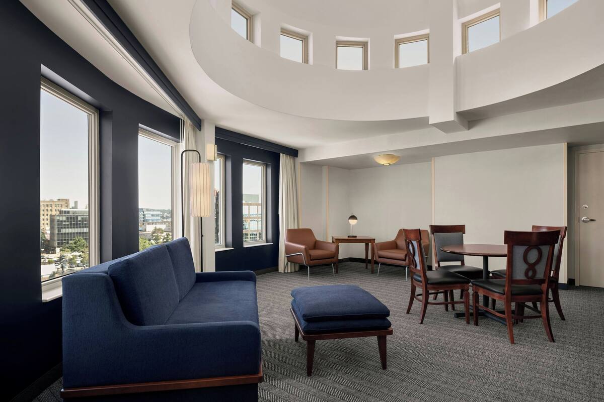 A modern hotel room with a curved ceiling, large windows offering city views, a blue sofa, a small table, and four wooden chairs.