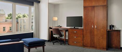 Habitación de hotel con un ventanal grande, un escritorio con computadora, un televisor, un armario y un área de descanso.