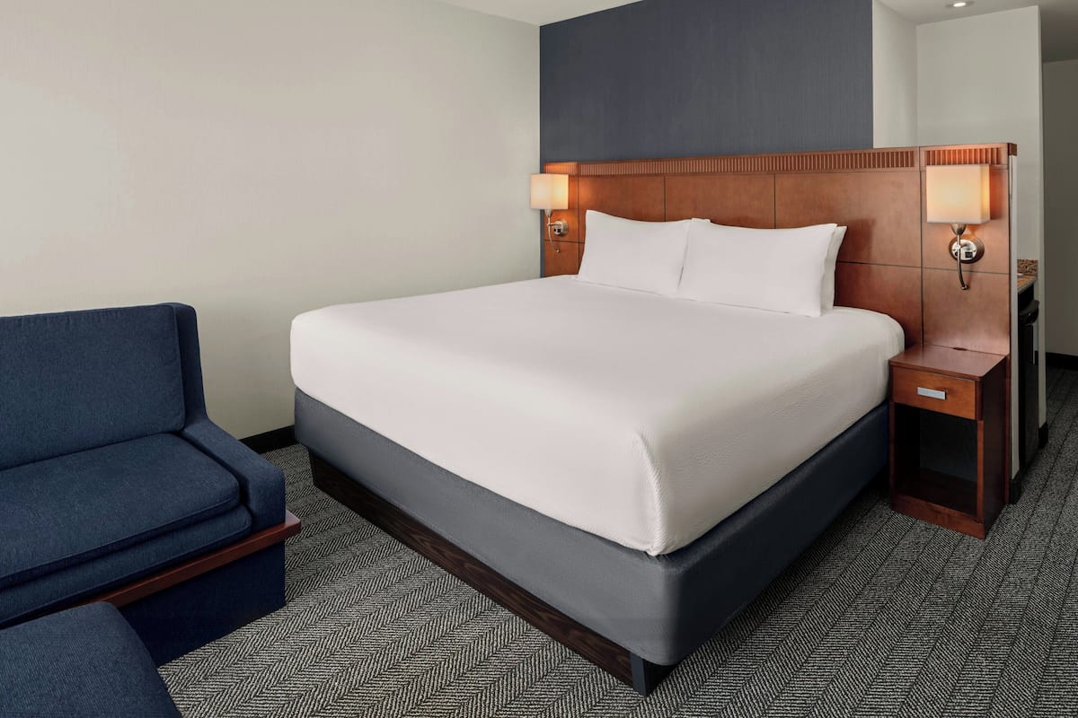 A hotel room with a large bed, two bedside tables with lamps, a blue armchair, and a herringbone-patterned carpet.