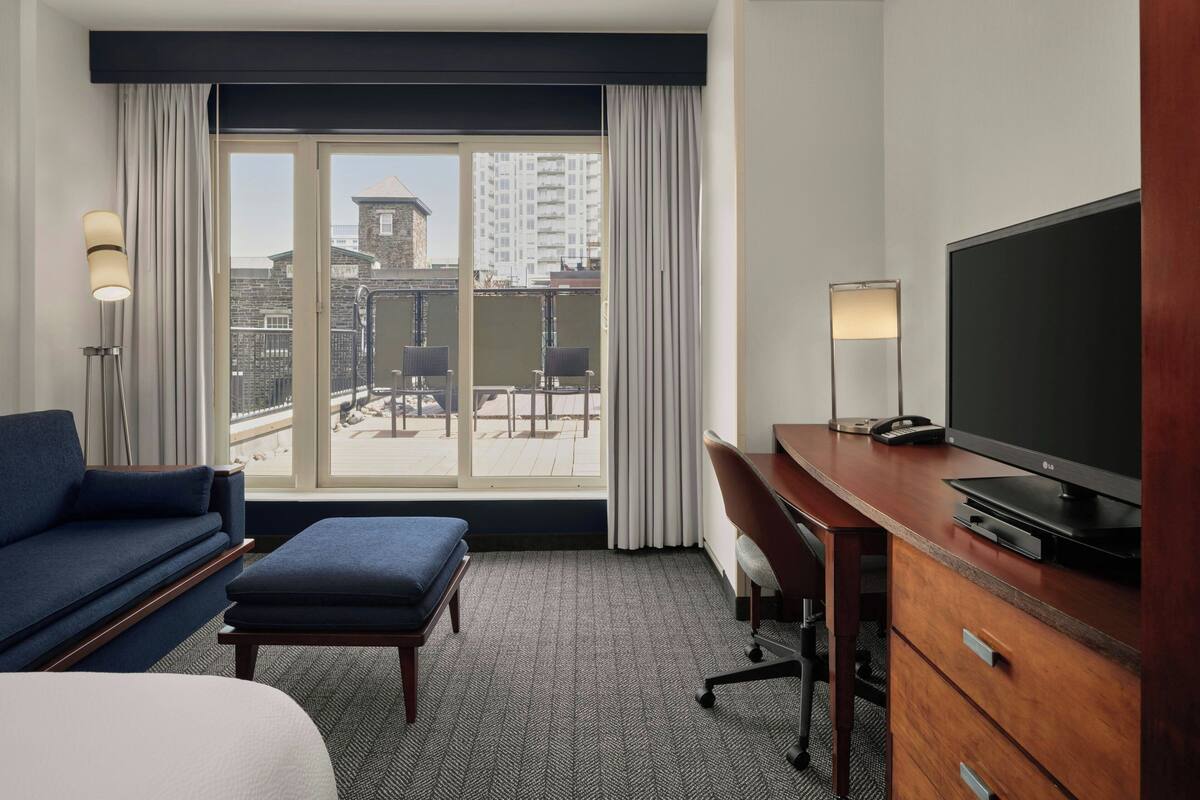 A hotel room with a large window, a sofa, a desk, a television, and a lamp.