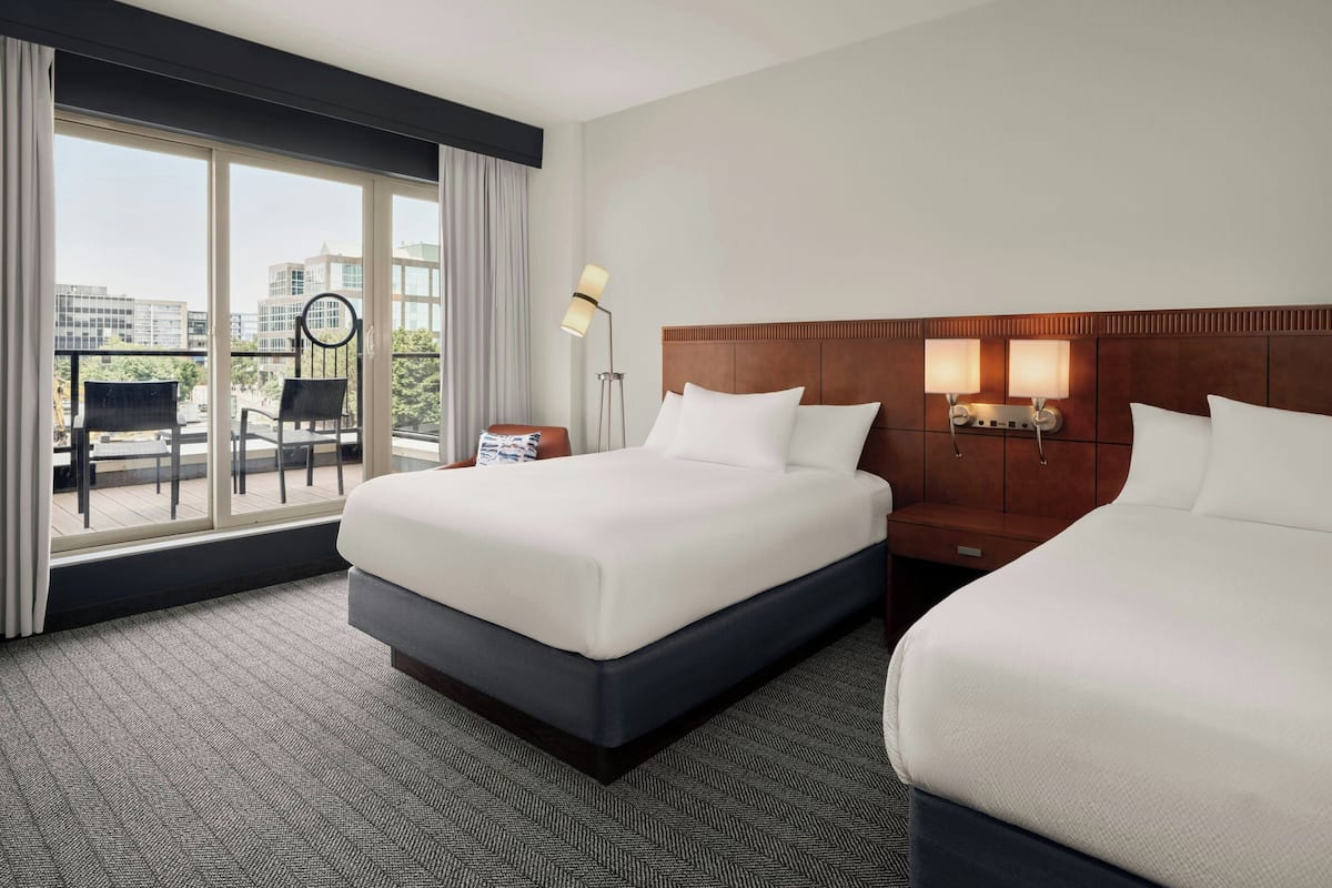 A hotel room with two beds, a large window with a view of buildings, a balcony with a chair, and a nightstand with a lamp.