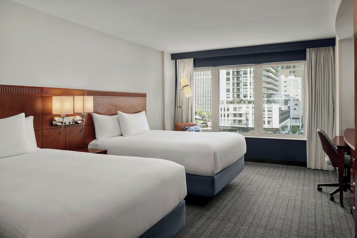 A hotel room with two beds, a desk, and a large window offering a city view.