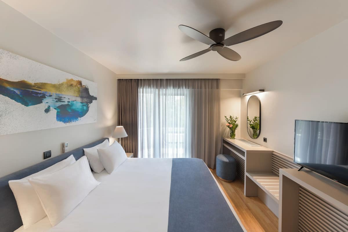 A modern hotel room with a large bed, a ceiling fan, a TV, and a desk with a mirror.