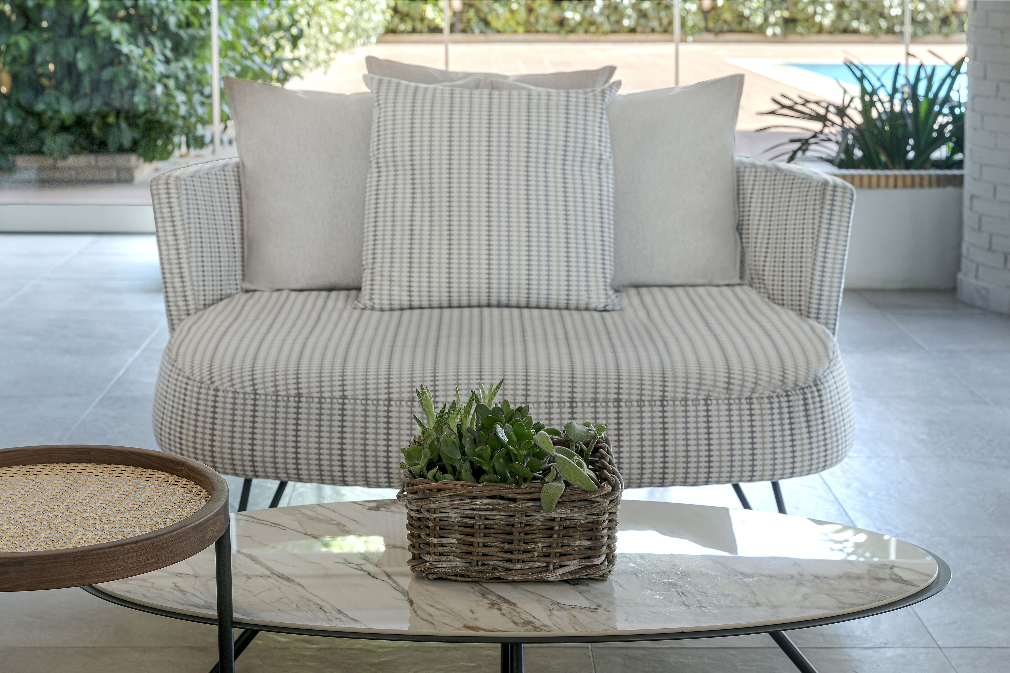 Un panier en osier contenant des plantes, posé sur une table en marbre, un canapé en osier avec des coussins et une chaise en osier avec un siège tressé.