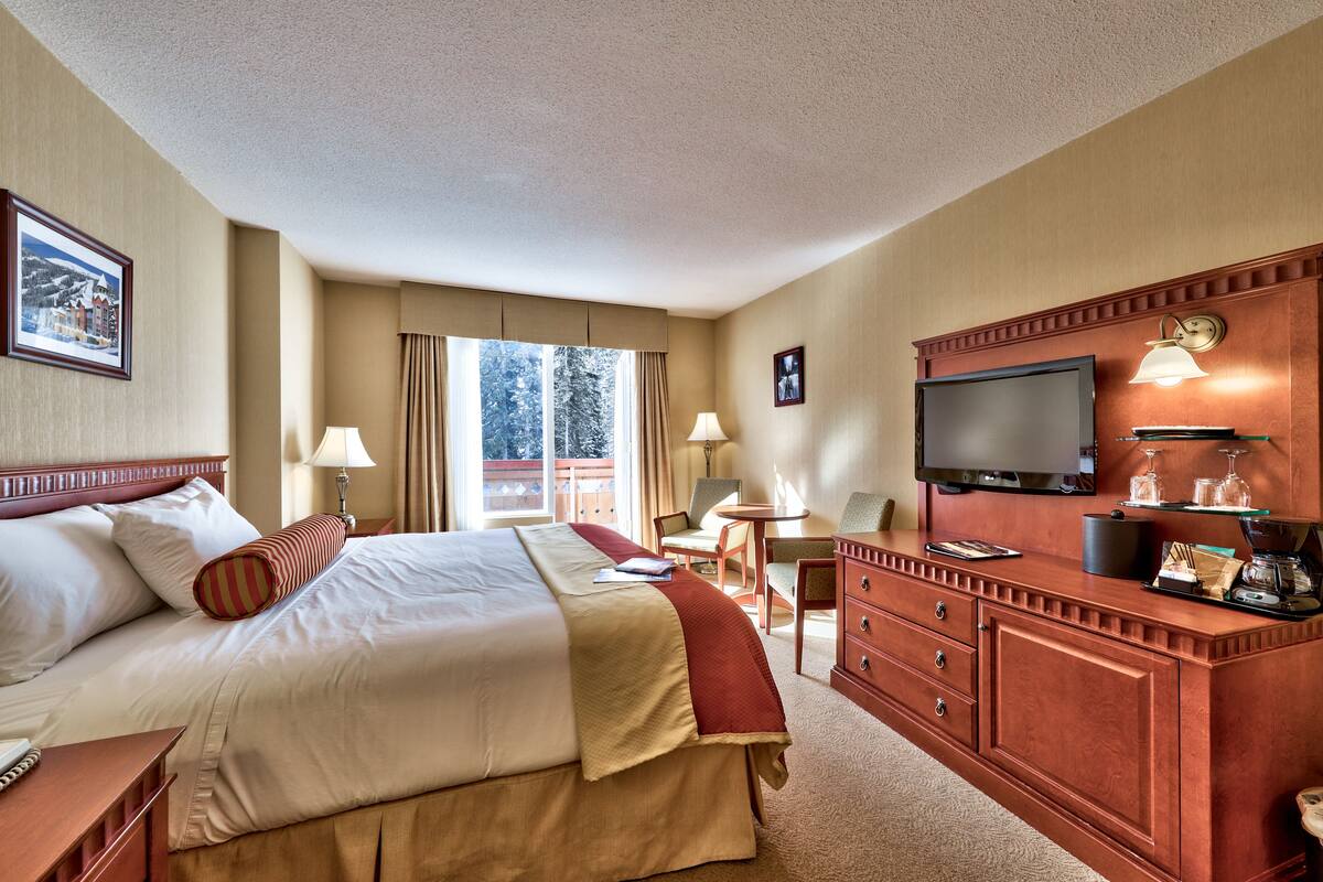 A hotel room with a large bed, a TV, a desk with a lamp, and a view of trees through the window.