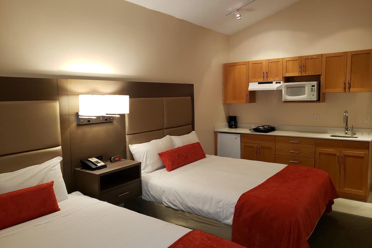 A hotel room with two beds, a small kitchenette, and a nightstand with a lamp and phone.