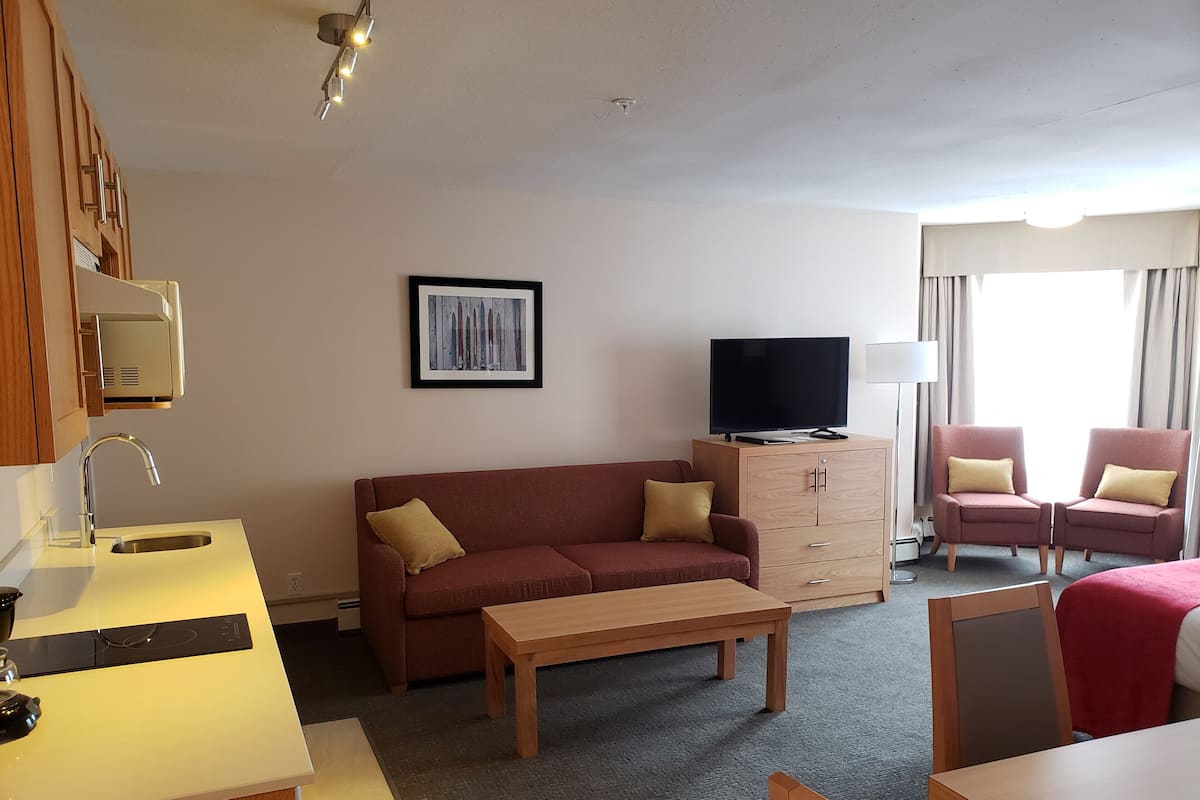 A hotel room with a sofa, two armchairs, a TV on a cabinet, a small table, and a kitchenette with a sink and stove.