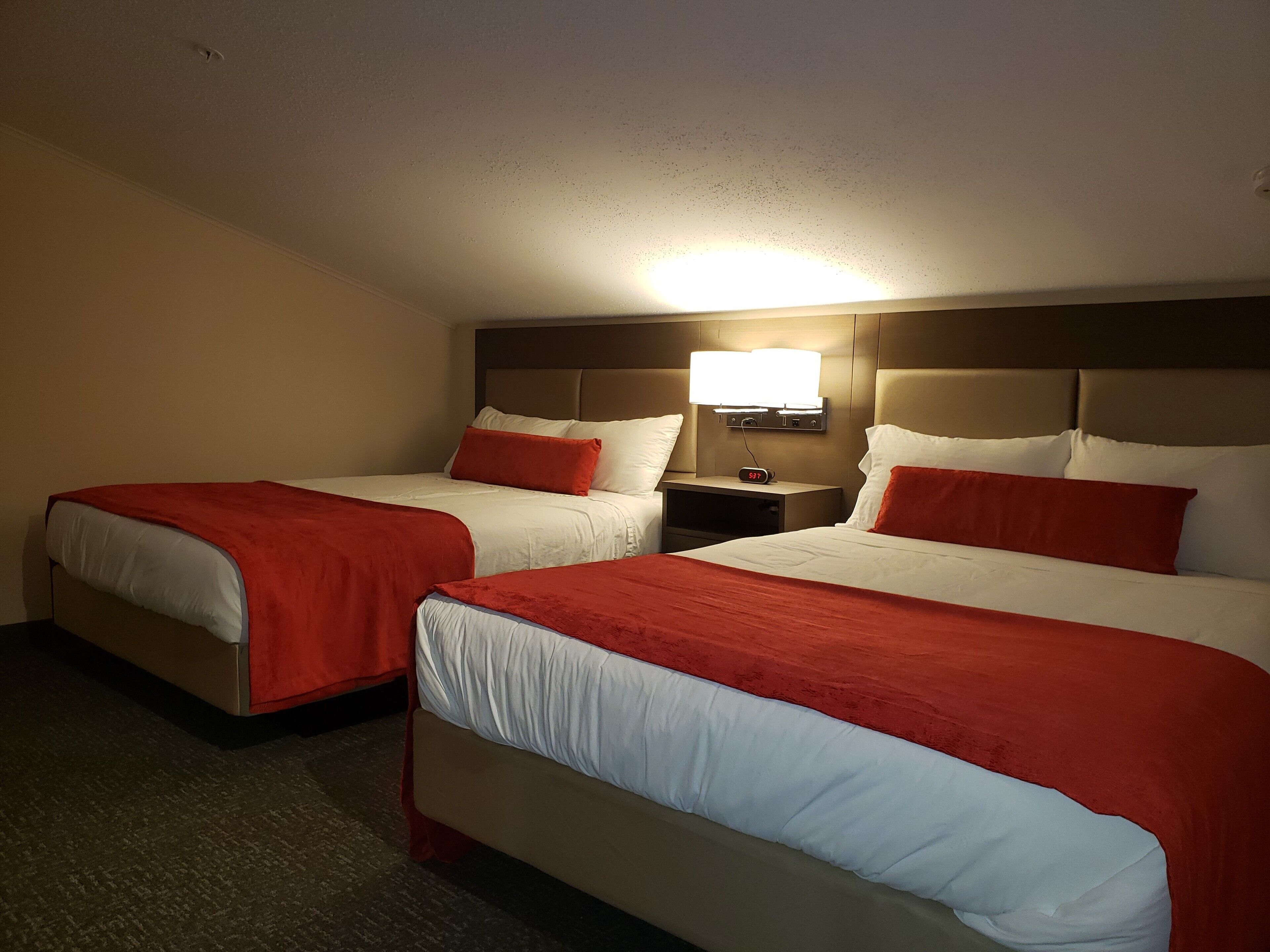 A hotel room with two beds, each with red and white bedding, a nightstand with a clock, and a wall-mounted light.