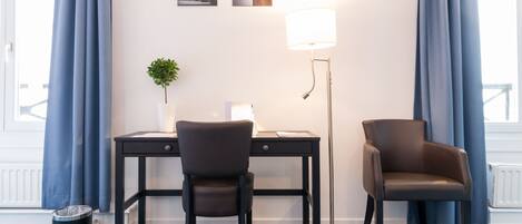 A room with a desk, two chairs, a floor lamp, a trash bin, and a potted plant.