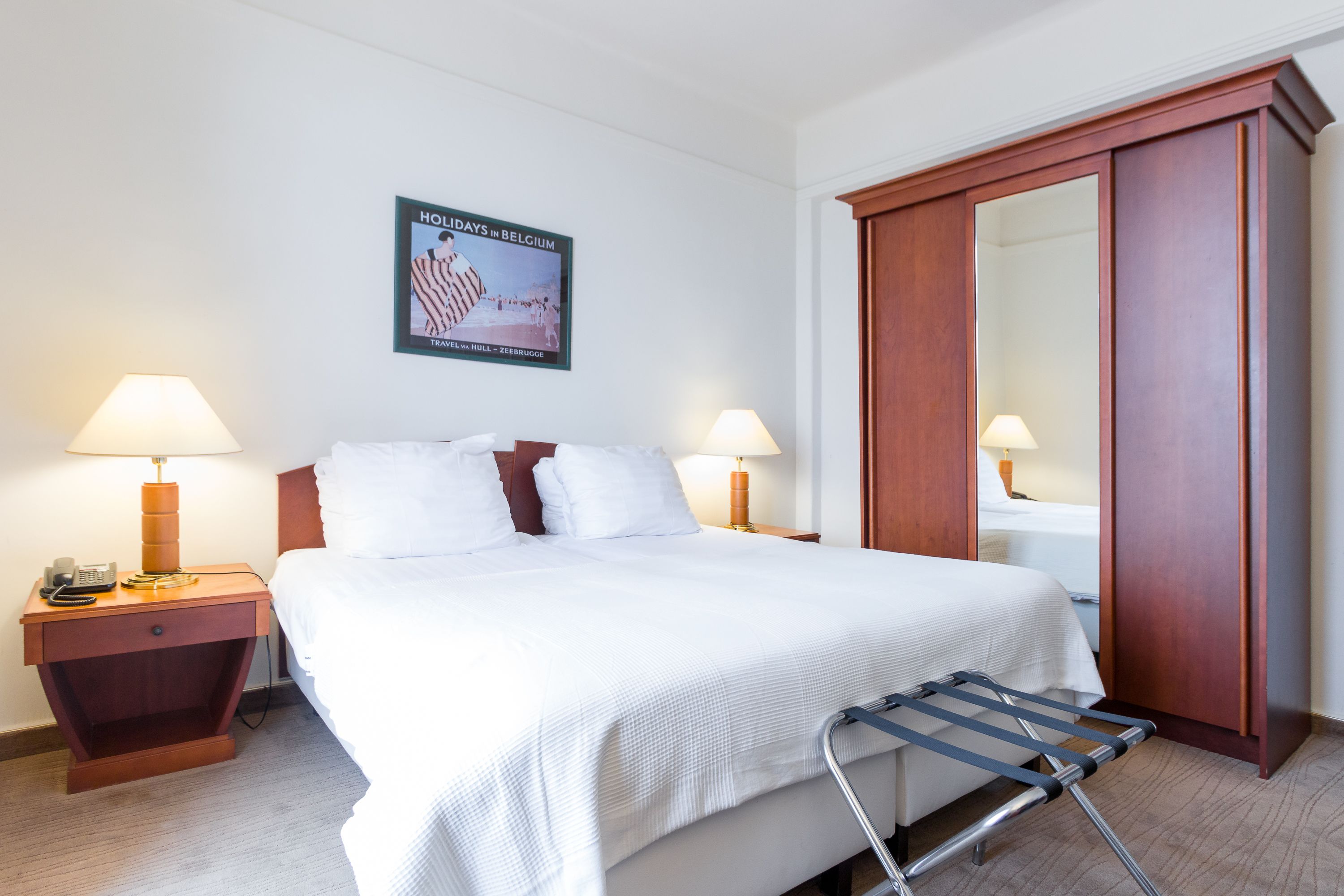 Een hotelkamer met een groot bed, twee nachtkastjes met lampen, een houten kledingkast en een poster aan de muur.