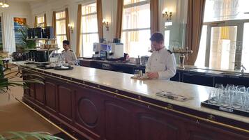 Een hotelbar met een marmeren toonbank, twee barmannen en uitzicht op de straat door grote ramen.